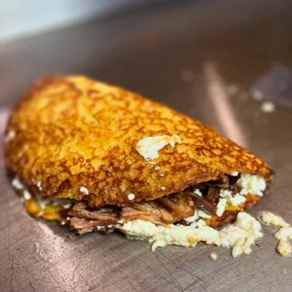 A golden-brown Chamo's Bagel & Grill arepa, filled with shredded meat and white cheese, on a metal surface.
