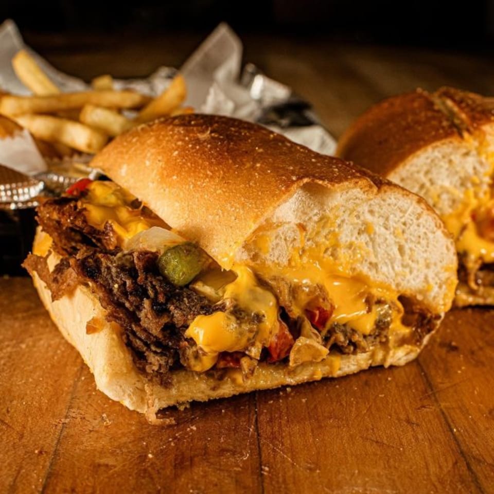 Cheesesteak sandwich with melted cheese, onions, and peppers, served with fries on a wooden table.