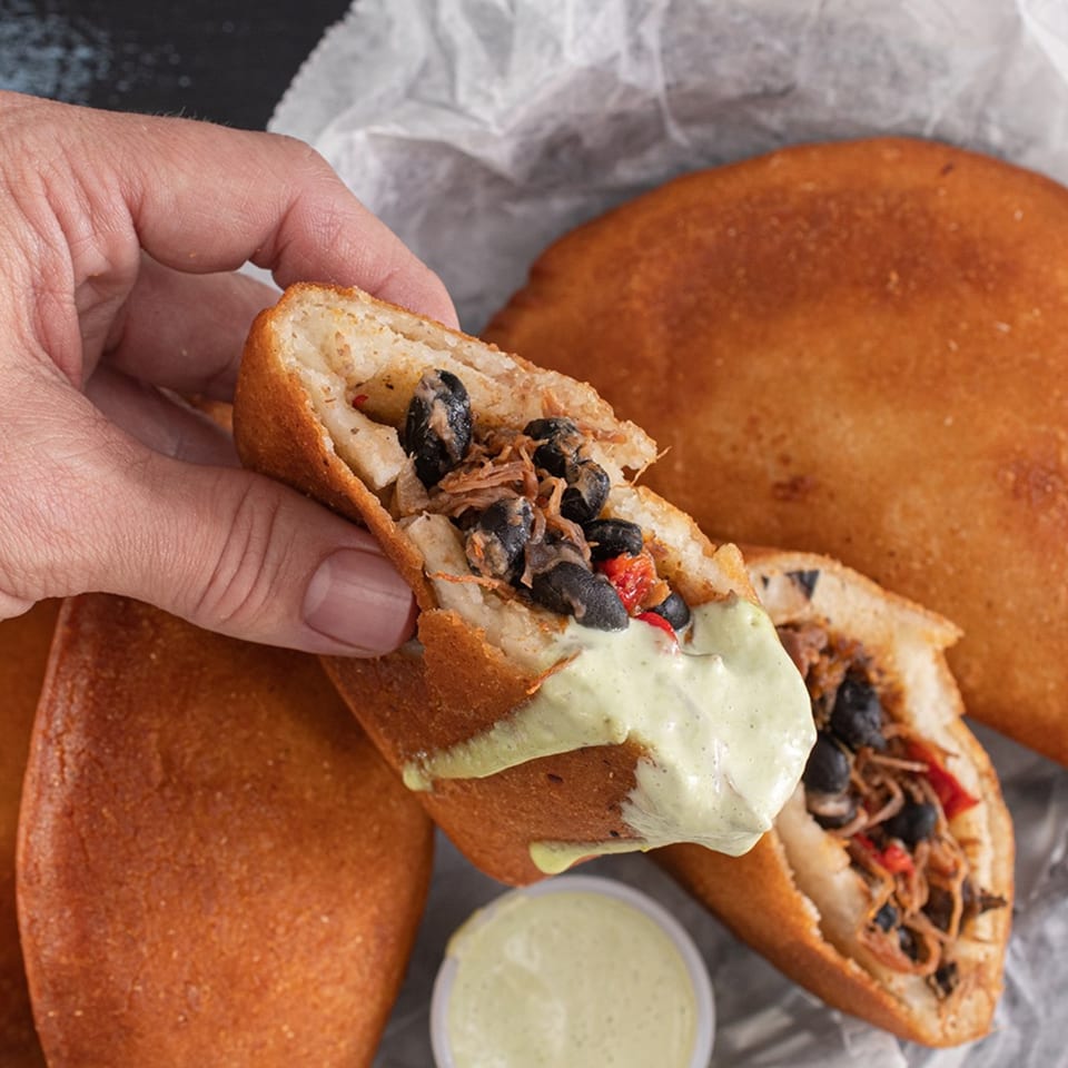 Hand holding a Chamo's Bagel & Grill empanada filled with black beans and meat, drizzled with sauce.